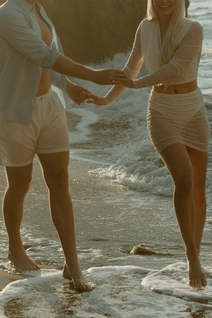 Engaged couple walking along the sandy shore of Malibu Beach during sunrise.