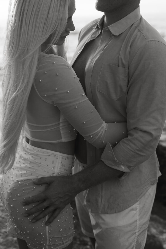 Close-up of engaged couple's loving embrace with the sunrise casting a warm glow on the coastline behind them.