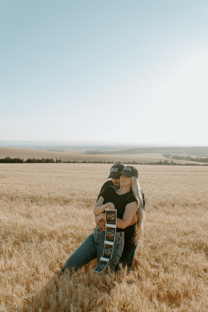 Pregnancy reveal photo shoot in a wheat field: Haley and Tate stand together amidst golden wheat stalks, holding ultrasound photos and a tiny onesie.