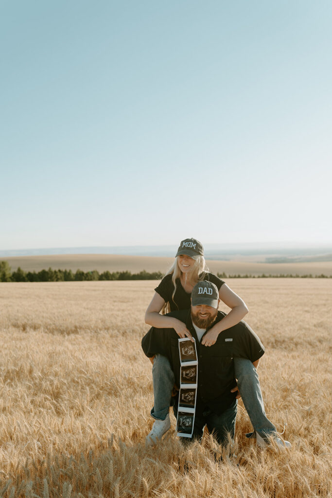 Pregnancy reveal photo shoot in a wheat field: Haley and Tate stand together amidst golden wheat stalks, holding ultrasound photos and a tiny onesie.