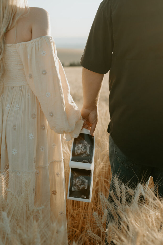 Pregnancy reveal photo shoot in a wheat field: Haley and Tate stand together amidst golden wheat stalks, holding ultrasound photos and a tiny onesie.