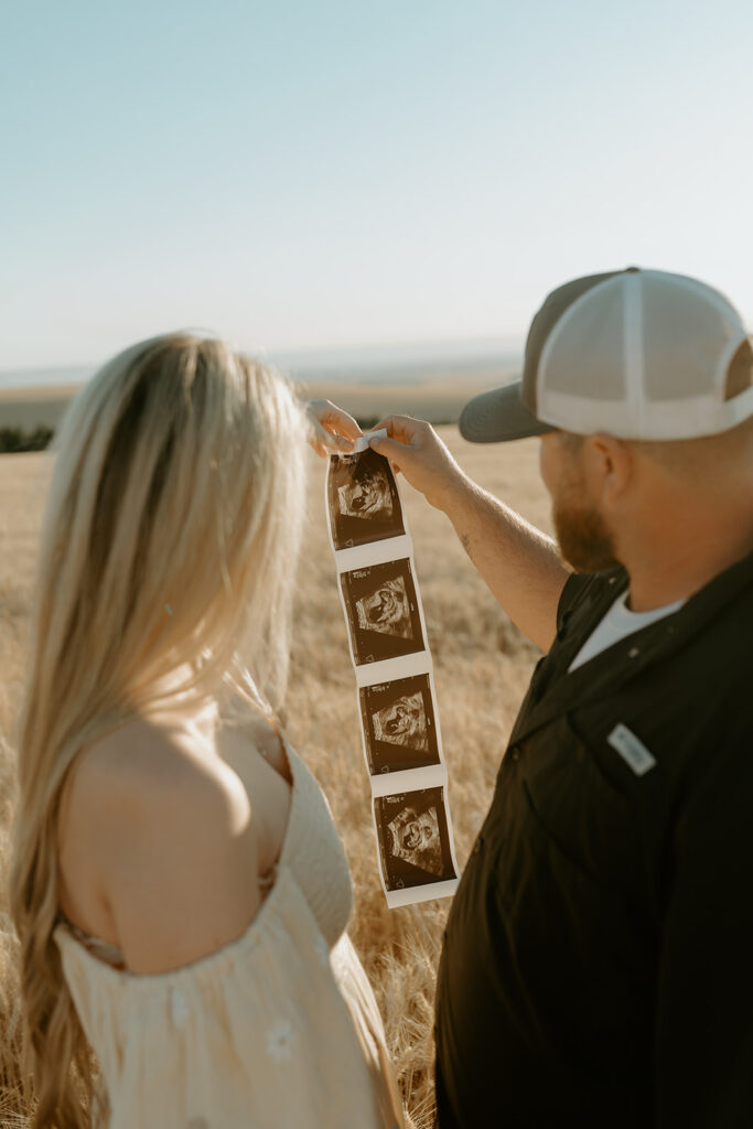 Pregnancy reveal photo shoot in a wheat field: Haley and Tate stand together amidst golden wheat stalks, holding ultrasound photos and a tiny onesie.