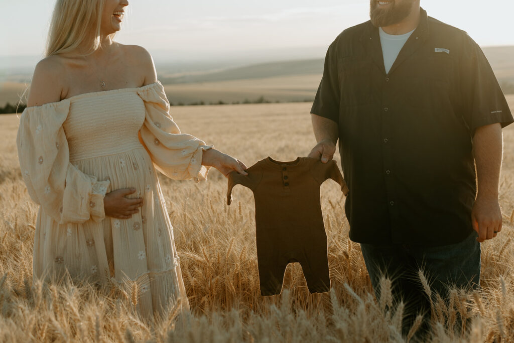 Pregnancy reveal photo shoot in a wheat field: Haley and Tate stand together amidst golden wheat stalks, holding ultrasound photos and a tiny onesie.