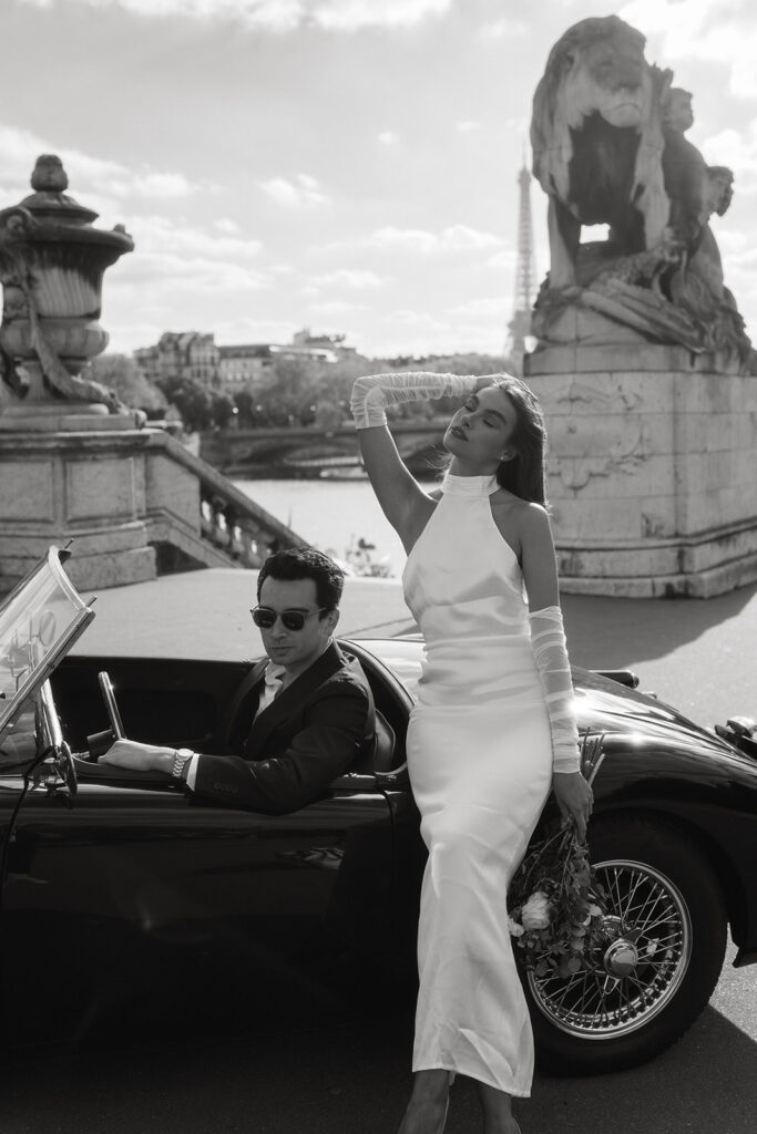 A couple poses in a vintage car parked in front of the Eiffel Tower in Paris, France, capturing a timeless and romantic moment in the city of love.