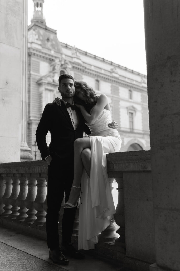 A couple embraces in front of the iconic Louvre Museum in Paris, France, with ornate architecture in the background.