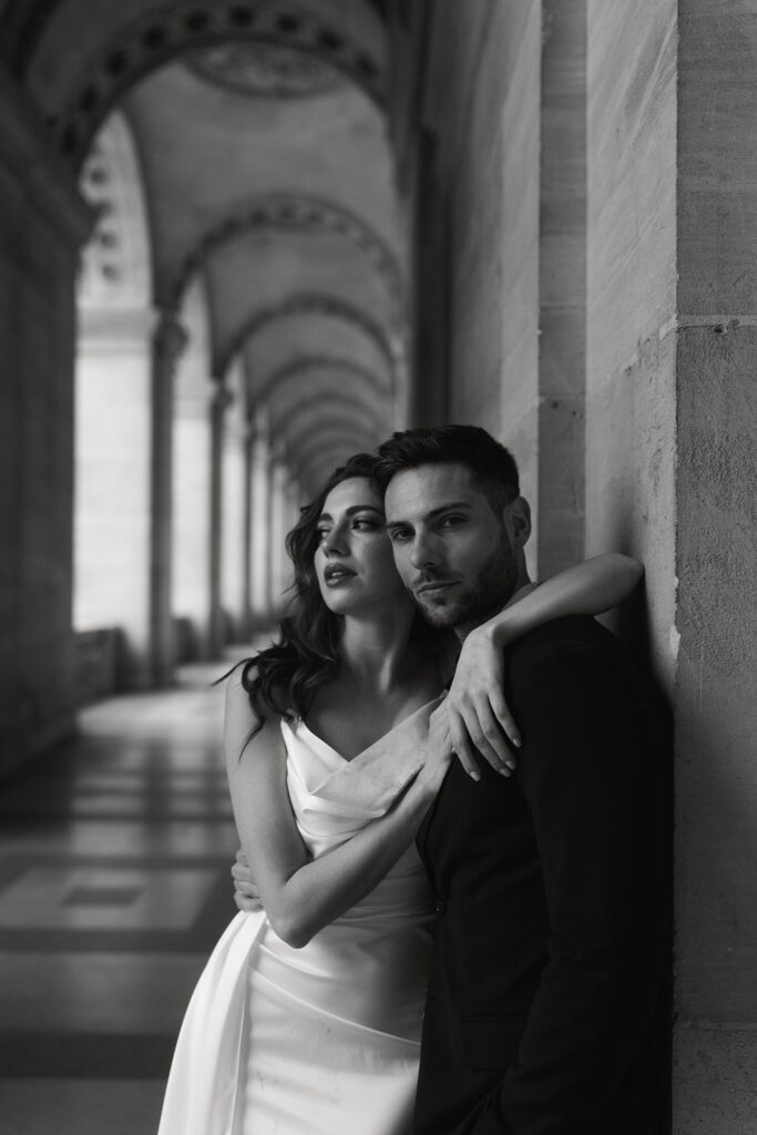 A couple embraces in front of the iconic Louvre Museum in Paris, France, with ornate architecture in the background.