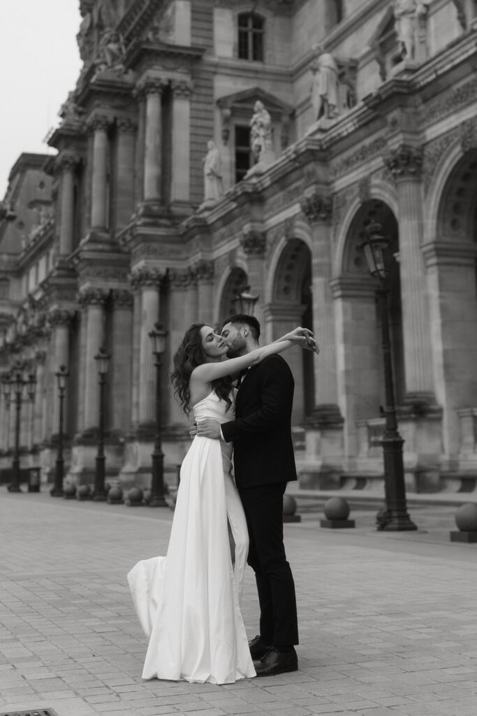 A couple embraces in front of the iconic Louvre Museum in Paris, France, with ornate architecture in the background.