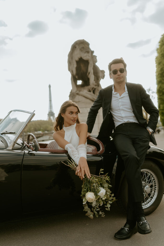 A couple poses in a vintage car parked in front of the Eiffel Tower in Paris, France, capturing a timeless and romantic moment in the city of love.