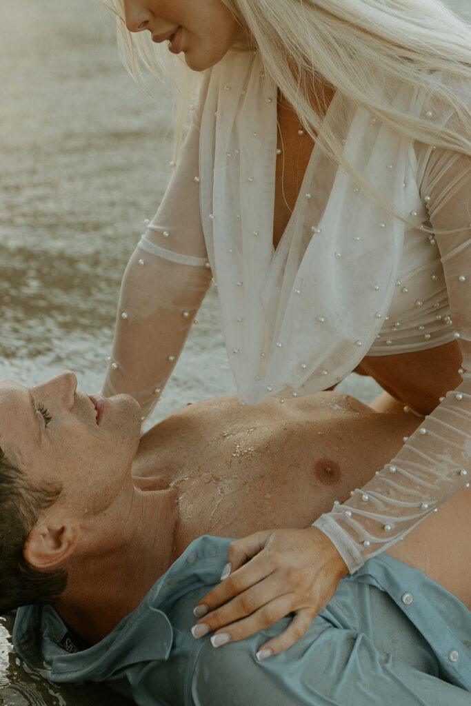 Close-up of engaged couple's loving embrace with the sunrise casting a warm glow on the coastline behind them.